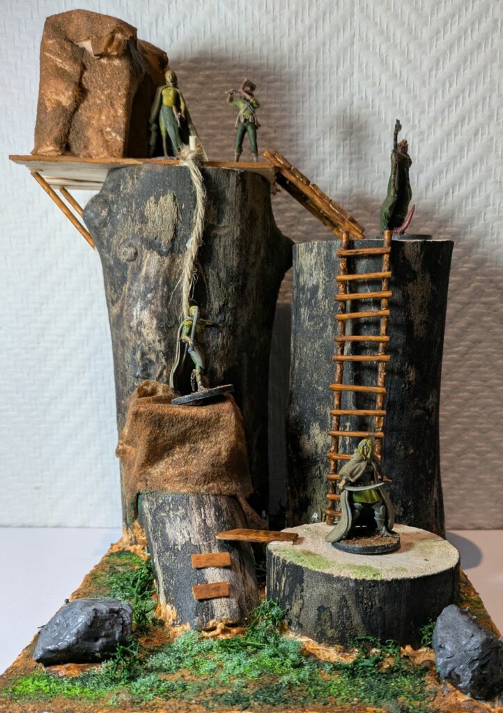 A terrain made of real wood, a dual wielding blond elf is standing on the lower level, another one is standing on an upper level, an elf with a bow is on the third elevation, and on top there are a hub and two elves playing music