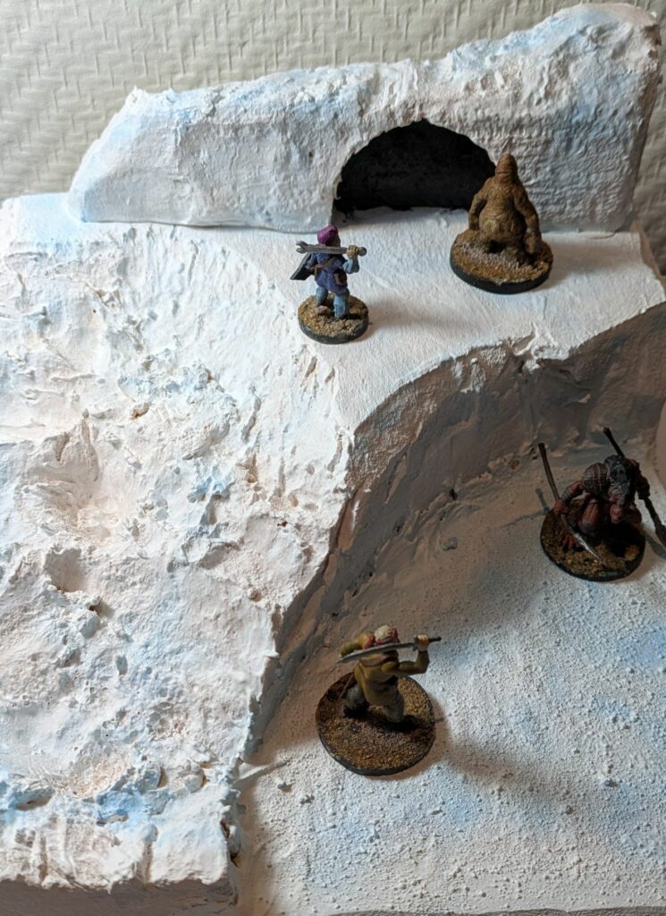 A snow and ice miniature terrain with a cliff and a grotto on the top. A gnome warrior is facing a brown yeti on the top, while a human warrior is facing a gnoll on the lower area.