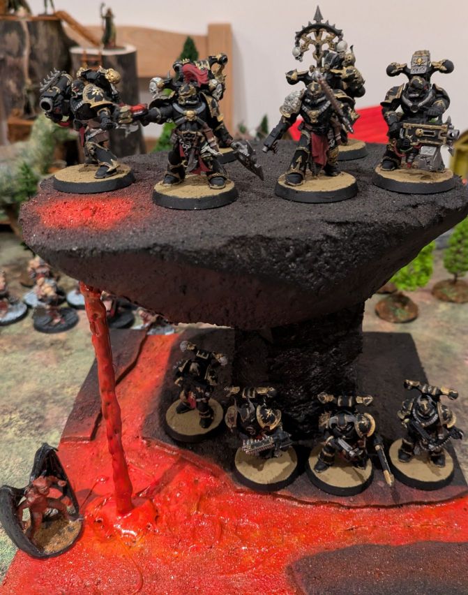 Ten Chaos Space Marines legionaries standing on a lava terrain, with a small red daemon in the lava