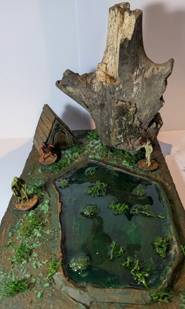 A miniature swamp made of isolation foam, a piece of dead wood, and resin. There is a green pond, a wooden shack and three monsters.