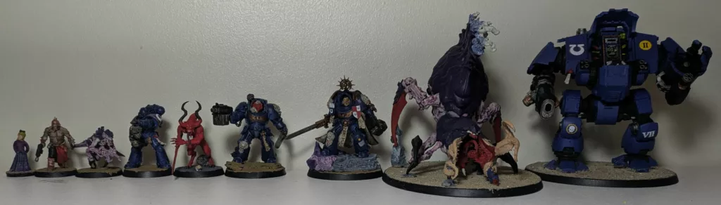 Nine Warhammer 40K miniatures side by side. A little girl, a Chaos Cultist, a Tyranid Termagaunt, an Ultramarine Infernus, a Chaos Daemon Bloodletter, an Ultramarine Terminator, a Captain in Terminator Armour, a Tyranid Psychophage and an Ultramarine Redemptor Dreadnought.